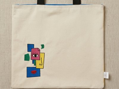 Trace Totes beige canvas tote featuring an embroidered abstract face graphic in blue, pink, yellow, and green with black webbing shoulder straps.