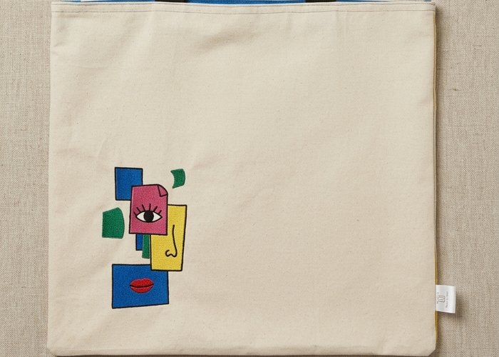 Trace Totes beige canvas tote featuring an embroidered abstract face graphic in blue, pink, yellow, and green with black webbing shoulder straps.