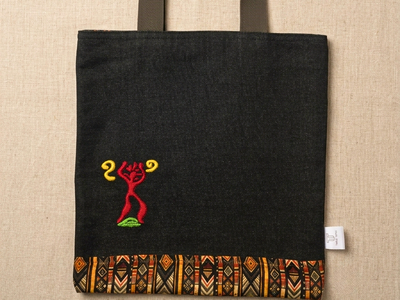 Trace Totes black denim tote bag with red and yellow tribal embroidered symbol, patterned fabric bottom border, and dark green woven handles laid flat on an oatmeal linen background.