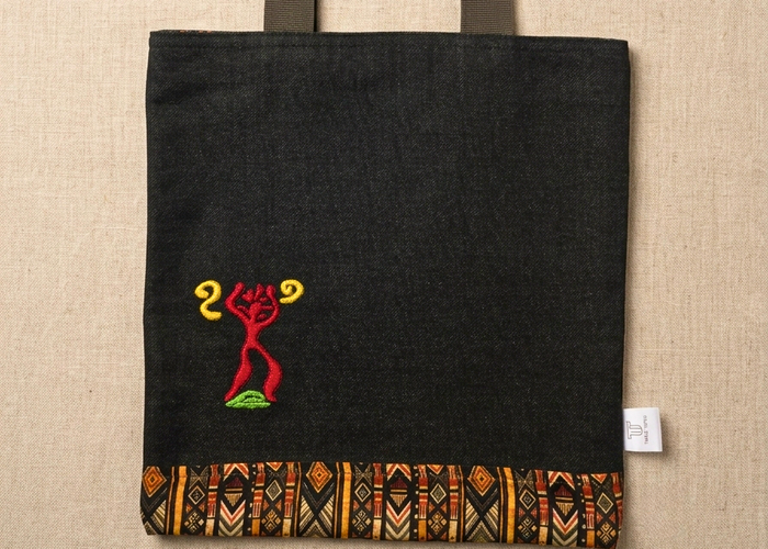 Trace Totes black denim tote bag with red and yellow tribal embroidered symbol, patterned fabric bottom border, and dark green woven handles laid flat on an oatmeal linen background.
