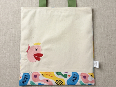 A Trace Totes canvas bag laid flat on oatmeal linen. Features an embroidered pink fish with a yellow crown, olive green straps, and a vibrant bottom panel with colorful abstract shapes in blue, yellow, pink, and green.