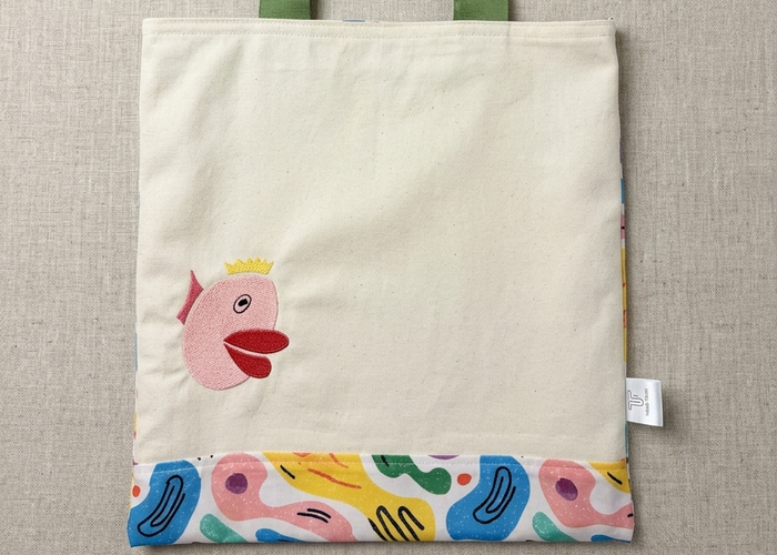 A Trace Totes canvas bag laid flat on oatmeal linen. Features an embroidered pink fish with a yellow crown, olive green straps, and a vibrant bottom panel with colorful abstract shapes in blue, yellow, pink, and green.