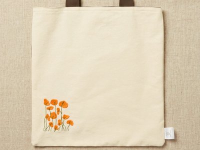 Trace Totes off-white canvas tote bag featuring embroidered orange poppies on the lower corner, dark brown webbing shoulder straps, and a brand tag, laid flat against an oatmeal linen background.