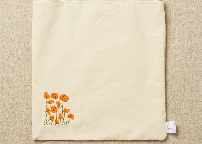 Trace Totes off-white canvas tote bag featuring embroidered orange poppies on the lower corner, dark brown webbing shoulder straps, and a brand tag, laid flat against an oatmeal linen background.