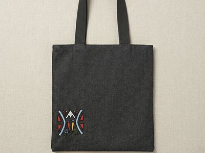 A front view of a black denim Trace Totes bag featuring a small, colorful geometric "funky face" embroidered in the bottom left corner with red, yellow, and blue accents. The bag has sturdy black shoulder straps and is set against a neutral beige backgrou