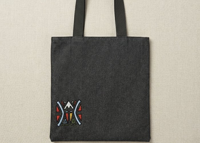 A front view of a black denim Trace Totes bag featuring a small, colorful geometric "funky face" embroidered in the bottom left corner with red, yellow, and blue accents. The bag has sturdy black shoulder straps and is set against a neutral beige backgrou