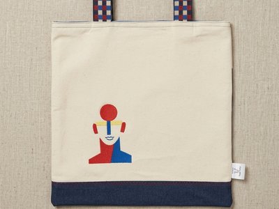 A Trace Totes beige canvas bag with a dark blue denim base and colorful abstract face graphic, featuring red and blue checkered woven straps against a linen background.