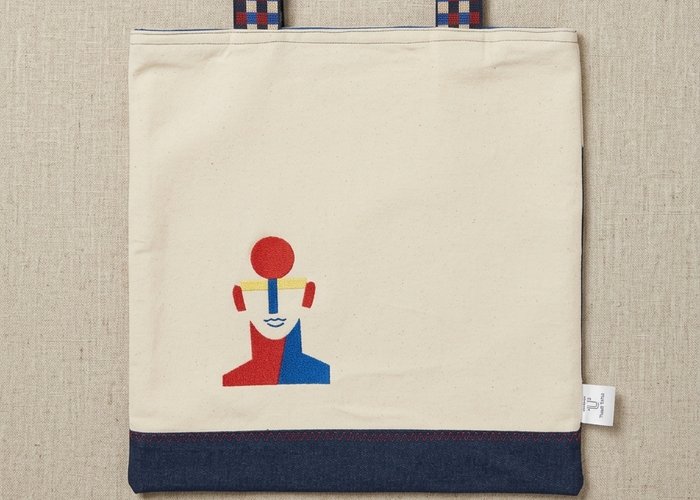 A Trace Totes beige canvas bag with a dark blue denim base and colorful abstract face graphic, featuring red and blue checkered woven straps against a linen background.