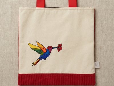 A Trace Totes beige canvas flat-lay tote bag featuring a vibrant, multi-colored geometric stained-glass style hummingbird embroidery at the bottom, red webbing handles, and a red corduroy bottom band, set against textured oatmeal linen.