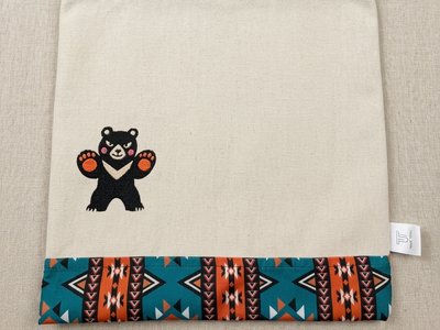 Trace Totes tote bag with a centered embroidered waving bear and a geometric pattern trim. Flat-laid on linen.