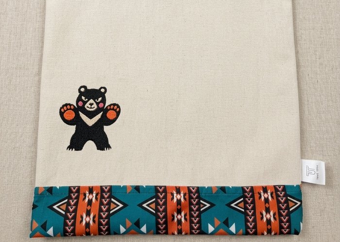 Trace Totes tote bag with a centered embroidered waving bear and a geometric pattern trim. Flat-laid on linen.