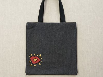 Square indigo denim tote bag featuring a black handle and a geometric red and yellow embroidered "kiss" in the lower corner.