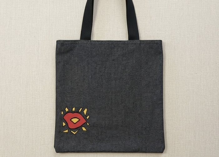 Square indigo denim tote bag featuring a black handle and a geometric red and yellow embroidered "kiss" in the lower corner.