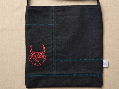 A black denim musette crossbody bag flat-laid by Trace Totes, featuring red graphic face embroidery, teal accent stitching, black webbing strap, and a white brand tag against an oatmeal linen fabric background.