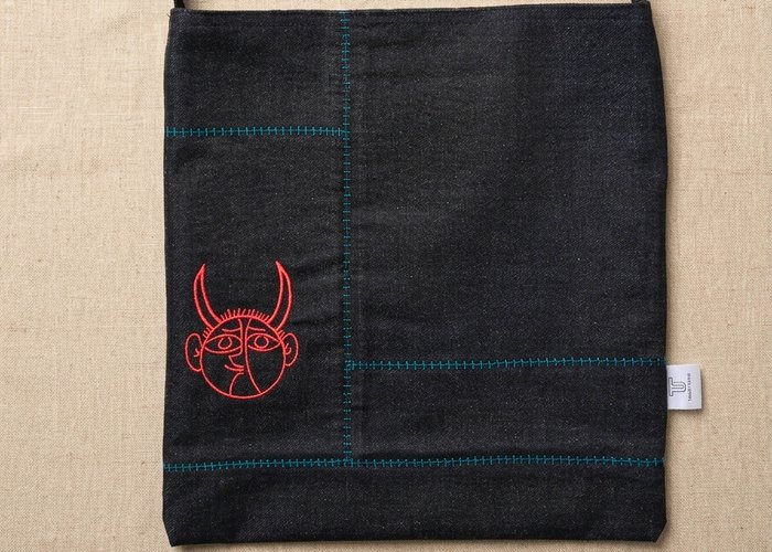 A black denim musette crossbody bag flat-laid by Trace Totes, featuring red graphic face embroidery, teal accent stitching, black webbing strap, and a white brand tag against an oatmeal linen fabric background.