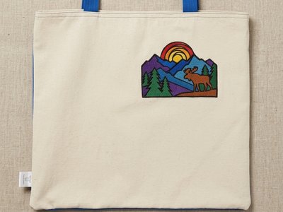 Trace Totes natural canvas tote bag featuring bright blue handles and a colorful embroidered mountain scene with a moose and sunset patch on the front right corner.