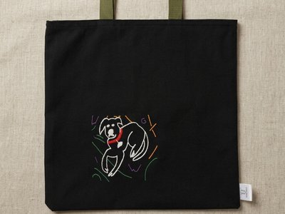 Trace Totes black canvas tote bag featuring an embroidered white line-art dog graphic on the lower left corner and olive green webbing shoulder straps, flat laid on an oatmeal linen fabric background.