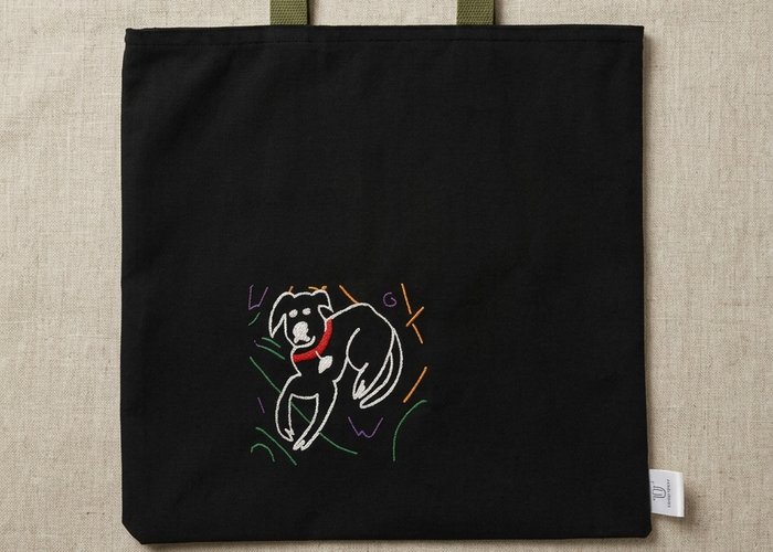 Trace Totes black canvas tote bag featuring an embroidered white line-art dog graphic on the lower left corner and olive green webbing shoulder straps, flat laid on an oatmeal linen fabric background.