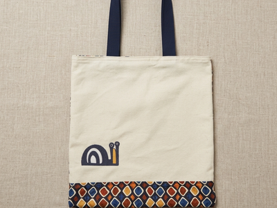 Flat-lay photo of a cream cotton Trace Totes tote bag with blue handles, a charming blue snail graphic on the front, and a vibrant, multi-colored geometric pattern border along the bottom. The bag rests centered on a high-quality oatmeal linen background