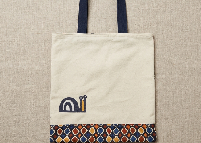 Flat-lay photo of a cream cotton Trace Totes tote bag with blue handles, a charming blue snail graphic on the front, and a vibrant, multi-colored geometric pattern border along the bottom. The bag rests centered on a high-quality oatmeal linen background