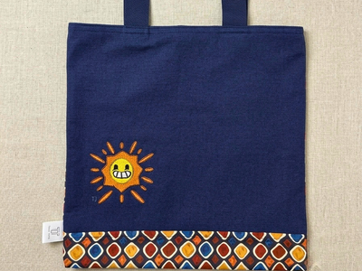 Premium navy canvas tote bag by Trace Totes featuring a friendly embroidered sun design, geometric patterned bottom trim, and sturdy navy straps laid flat on oatmeal linen.