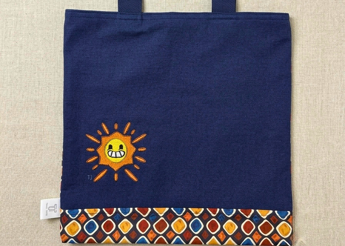 Premium navy canvas tote bag by Trace Totes featuring a friendly embroidered sun design, geometric patterned bottom trim, and sturdy navy straps laid flat on oatmeal linen.