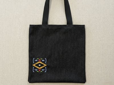 High-resolution flat lay of a black denim tote bag featuring a vibrant, multi-colored geometric embroidered design on the bottom left. The bag includes sturdy black canvas straps and is centered on a neutral, textured oatmeal linen background.