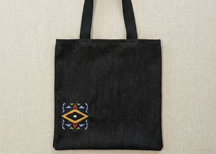 High-resolution flat lay of a black denim tote bag featuring a vibrant, multi-colored geometric embroidered design on the bottom left. The bag includes sturdy black canvas straps and is centered on a neutral, textured oatmeal linen background.