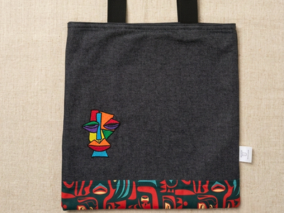 Charcoal canvas tote bag by Trace Totes featuring a colorful cubist face embroidery and red-patterned trim, laid flat on an oatmeal linen fabric.