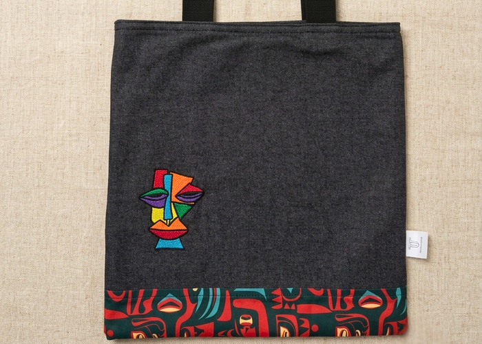 Charcoal canvas tote bag by Trace Totes featuring a colorful cubist face embroidery and red-patterned trim, laid flat on an oatmeal linen fabric.