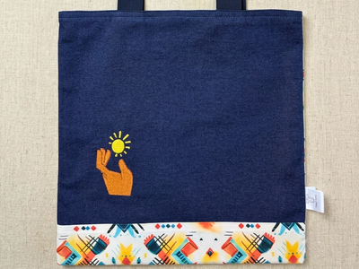 High-resolution product image of a dark blue denim tote bag by Trace Totes laid flat on an oatmeal linen surface. Features a small embroidered hand holding a sun icon and a colorful geometric patterned border along the bottom hem.