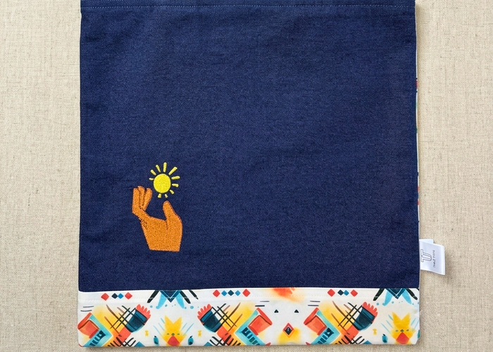 High-resolution product image of a dark blue denim tote bag by Trace Totes laid flat on an oatmeal linen surface. Features a small embroidered hand holding a sun icon and a colorful geometric patterned border along the bottom hem.